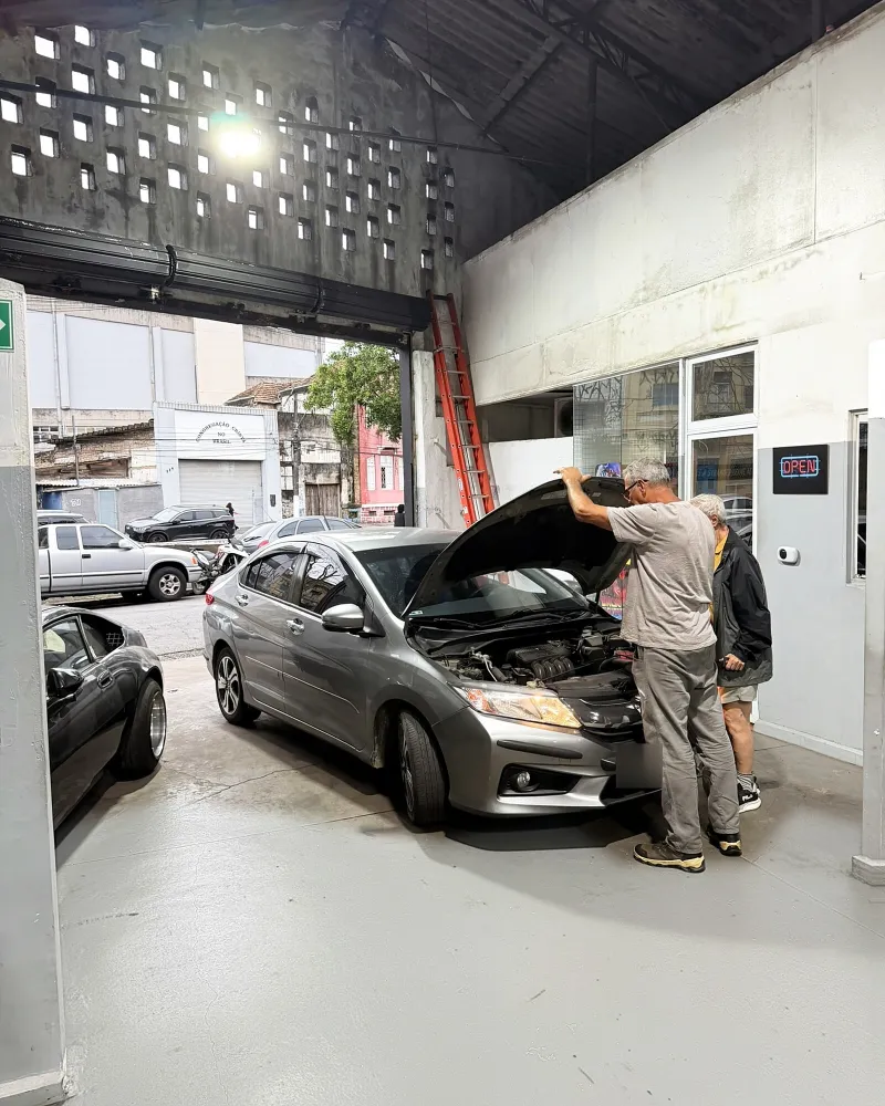 Mecânico abre o capô de um sedã prata na entrada da oficina, ao lado do cliente