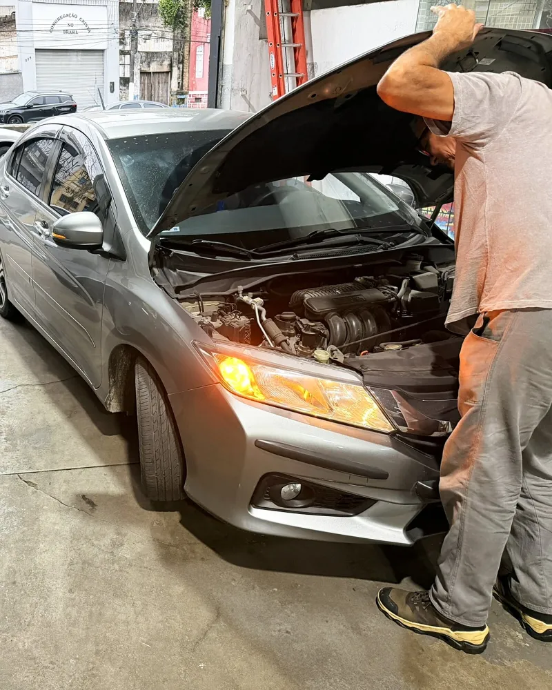 Mecânico inclinado sobre o motor de um carro prata, com o farol aceso