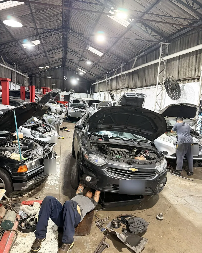 Galpão da oficina com dois mecânicos trabalhando, um deitado sob um carro e outro no motor de outro veículo